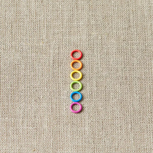 Six colors of Cocoknits Small Stitch Markers arranged in a neat vertical row on natural linen