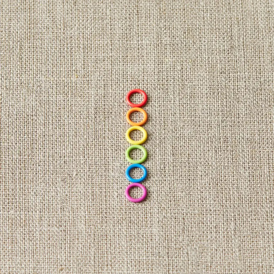 Six colors of Cocoknits Small Stitch Markers arranged in a neat vertical row on natural linen