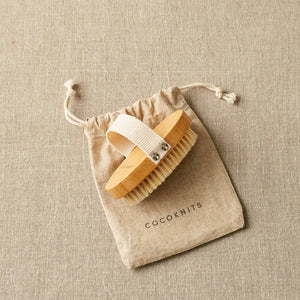 Cocoknits Sweater Care Brush. Wooden brush with its hand strap beside the drawstring bag.