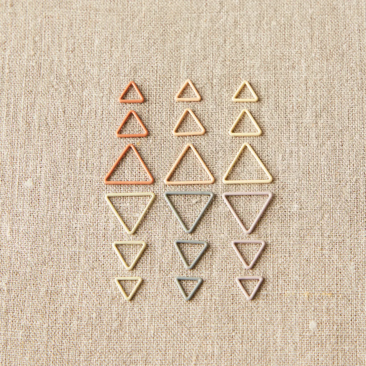 Cocoknits Triangle Stitch Markers in Earth Tones arranged by size and color on a natural linen background