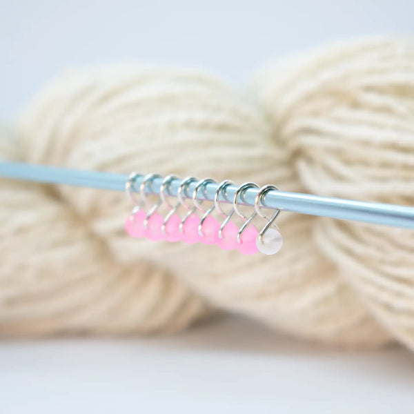 Extra Small Snag-Free Stitch Markers – Silver | Frosted Cookie. Pink stitch markers attached to a knitting needle beside cream yarn.