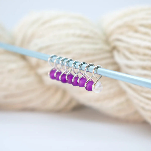 Extra Small Snag-Free Stitch Markers – Silver | Grape Slush. Purple stitch markers attached to a knitting needle beside cream yarn.
