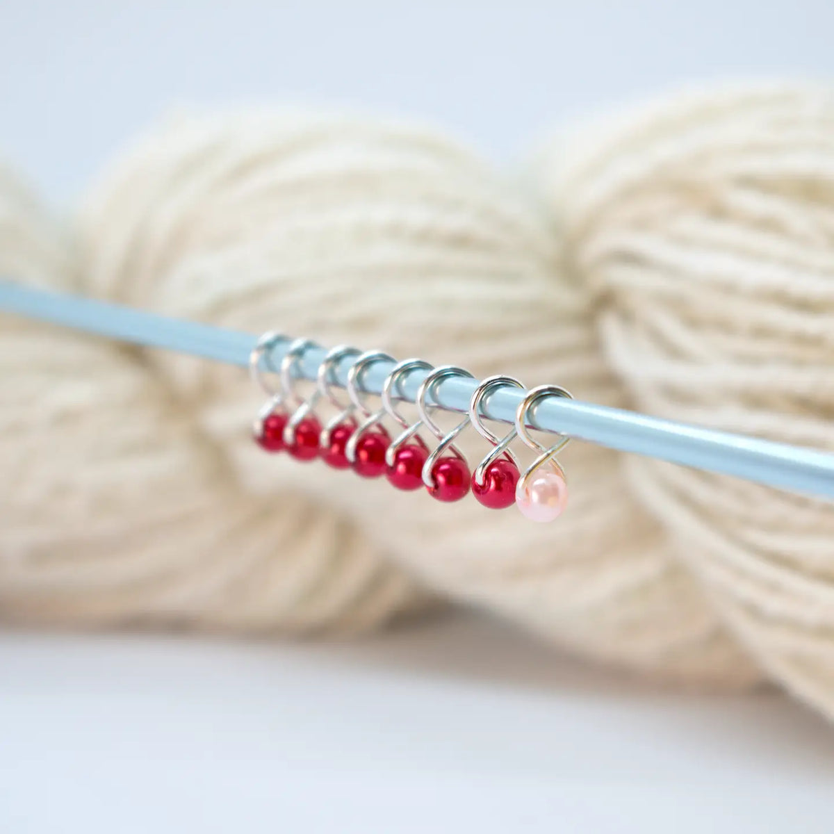 Extra Small Snag-Free Stitch Markers – Silver | Ruby. Red stitch markers attached to a knitting needle beside cream yarn.