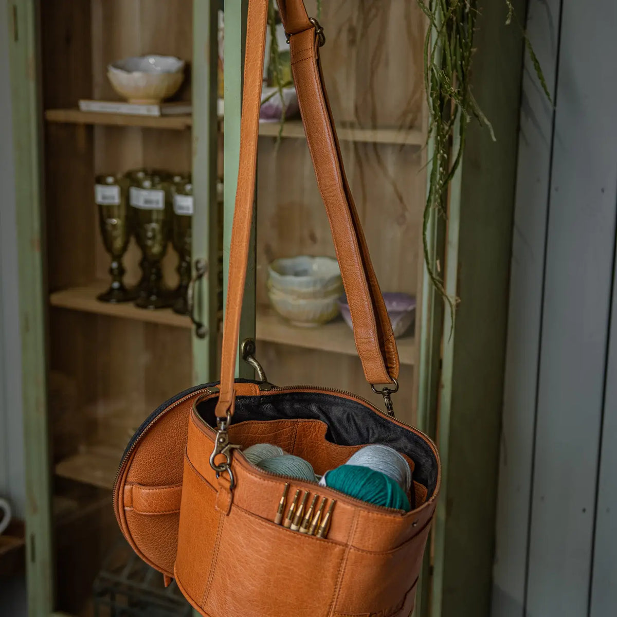 Muud Enturn Leather Project Bag | Black in Whisky. Open project bag holding yarn and knitting tools beside a wooden chair.